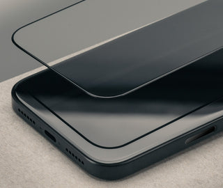A close-up of a black smartphone with a transparent tempered glass screen protector partially lifted over the top surface, set on a light neutral textured surface.